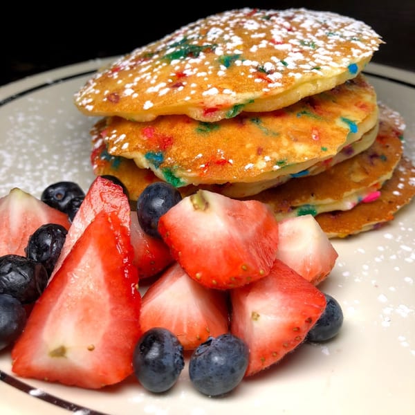 Stack of confetti pancakes dusted with powdered sugar, alongside fresh strawberries and blueberries.
