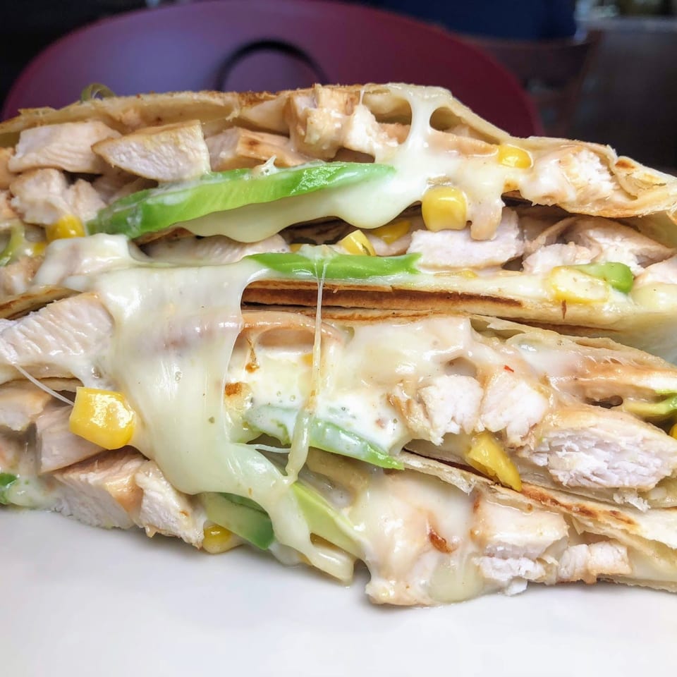 Close-up of a chicken quesadilla with melted cheese, corn, and avocado slices on a white plate.